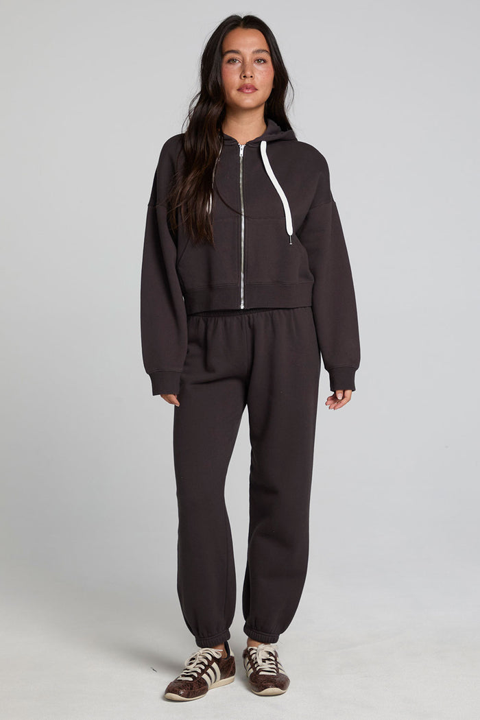 Full body front view of a woman wearing the Chaser  Abilene Dark Heather Grey Zip Up on a neutral background with matching  track pants