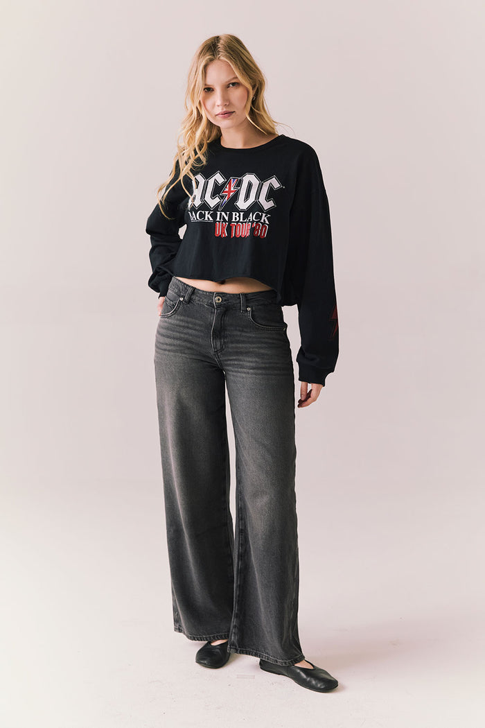 Full body view of model wearing AC/DC UK Tour ’80 Tee in True Black with cropped long-sleeve fit and vintage tour poster graphic paired with jeans on plain background