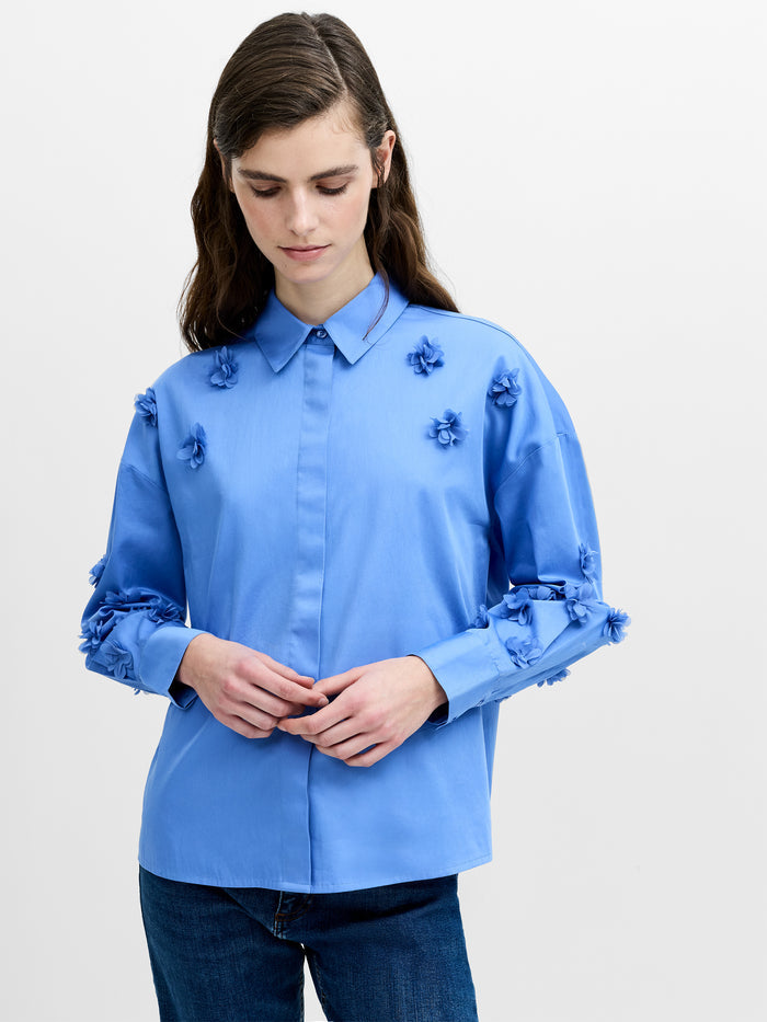 A woman wearing a blue cotton shirt with a 3D floral print, long sleeves, button down front, shirt collar, pleated back, dropped shoulders and a relaxed boxy fit.