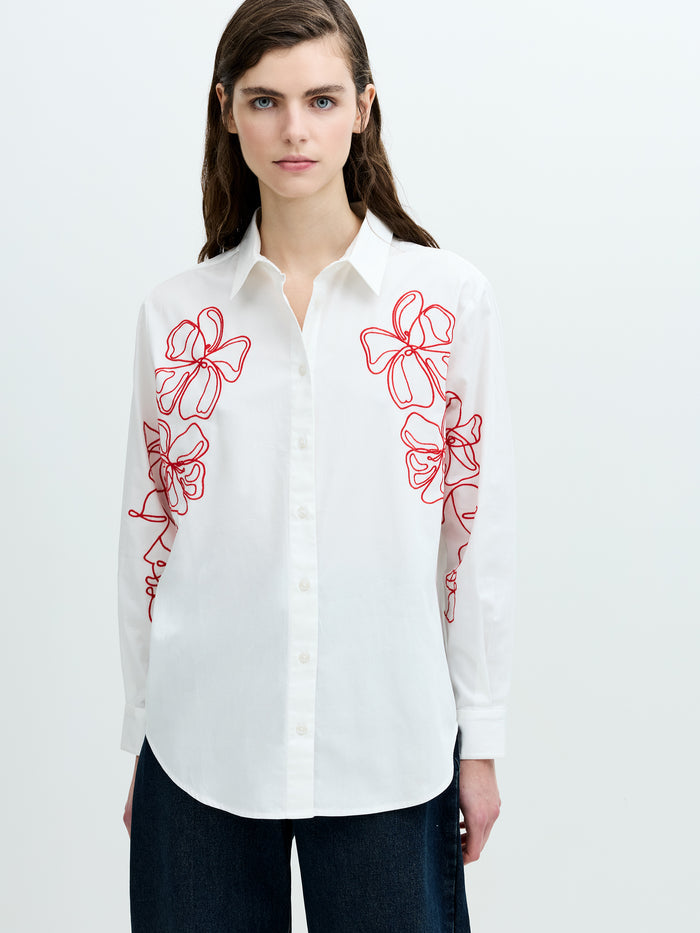 A woman wearing a white long sleeved shirt with red floral embroidery, a pointed collar and a back box pleat.