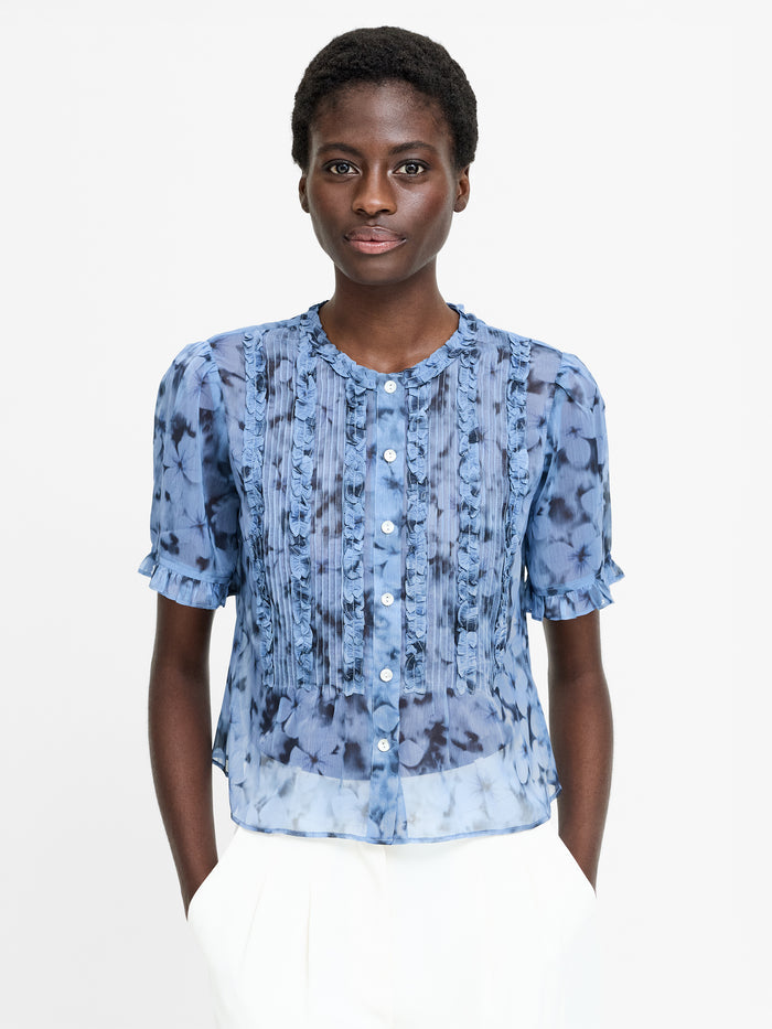 A woman wearing a short sleeved semi sheer floral print ruffle top with front buttons and a round neckline.