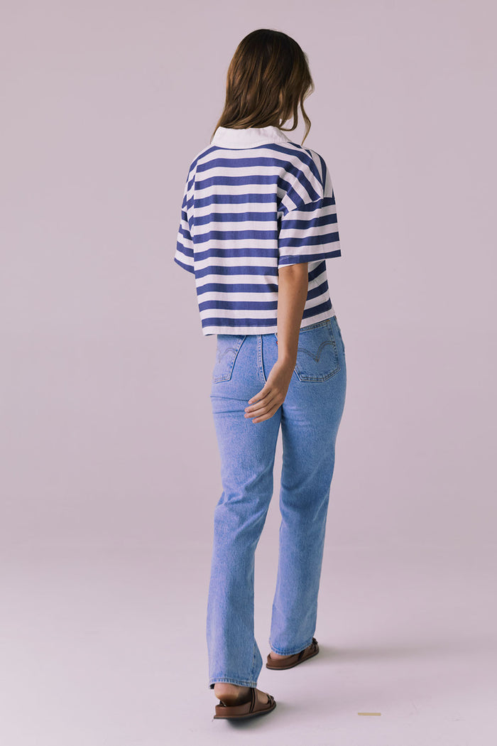 Back view of a woman wearing the Chaser Antoni Castaway Stripe Tee styled with high-waisted light-wash jeans and sandals in a minimalist studio setting