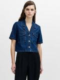 A woman wearing a short sleeved blue denim shirt with a revere collar, chest pockets, a boxy cropped fit and a button through fastening.