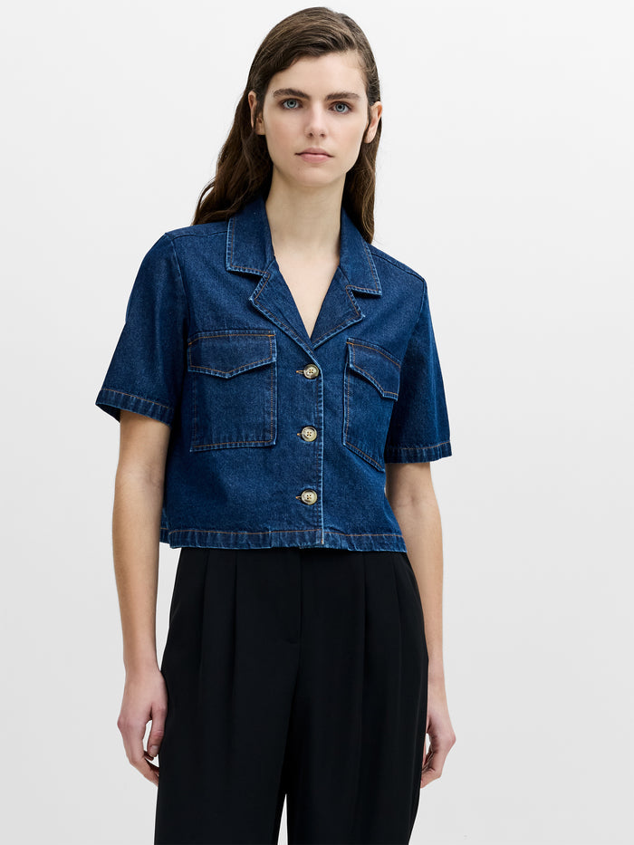 A woman wearing a short sleeved blue denim shirt with a revere collar, chest pockets, a boxy cropped fit and a button through fastening.