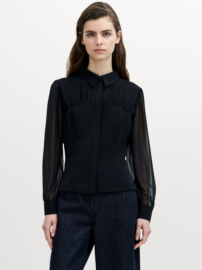 A woman wearing a black corset shirt with a button through fastening, sheer chest and long cuffed sleeves.