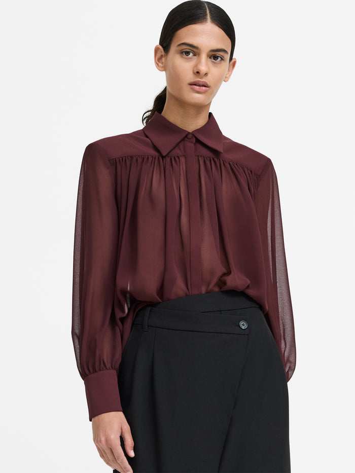 A woman wearing a wide shoulder burgundy shirt with long bishop sleeves, a button through fastening and a relaxed fit.