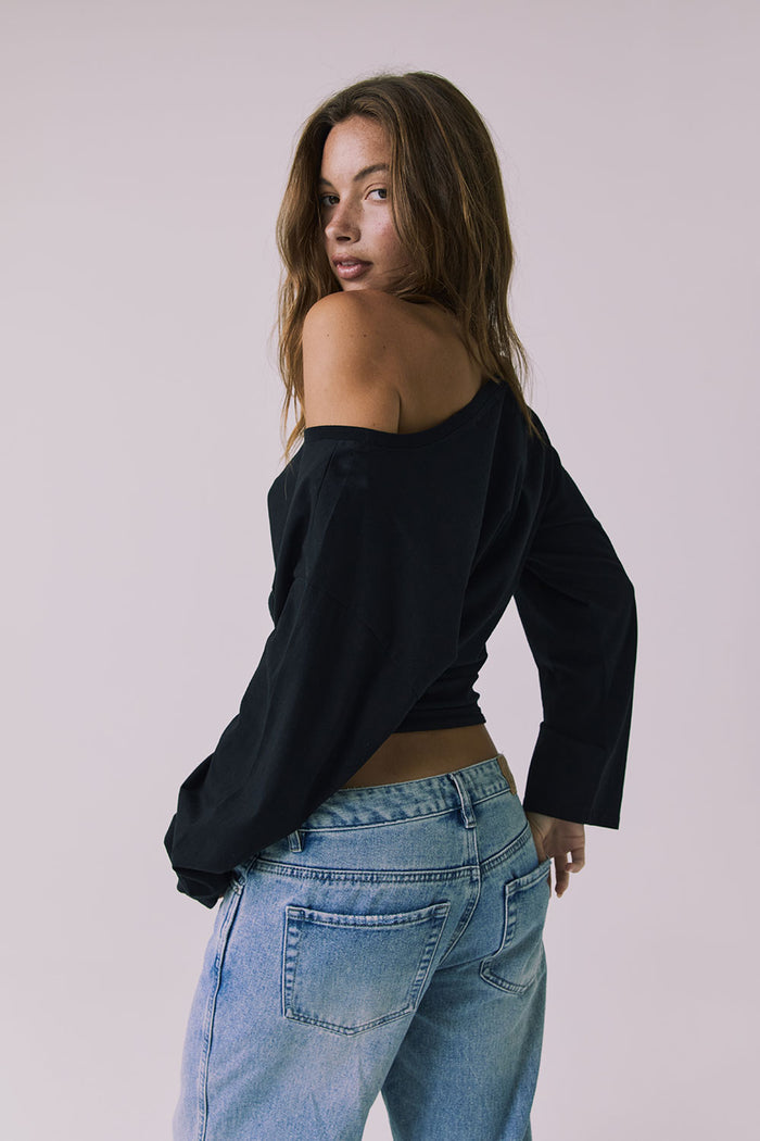 Back view of a woman wearing Chaser off-shoulder Ash Black Long Sleeve and blue jeans against a plain background
