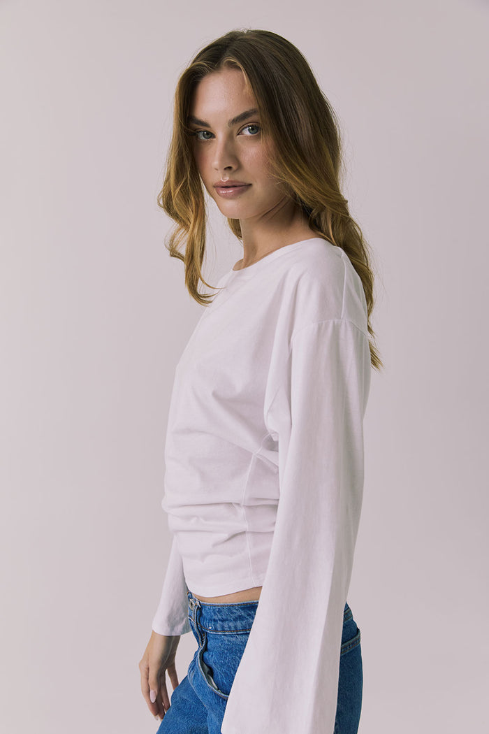 Side view of a woman wearing Chaser's Ash White Long Sleeve and blue jeans against a plain background