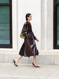 A woman wearing a burgundy belted  PU trench coat with wide lapels, side pockets and long sleeves.