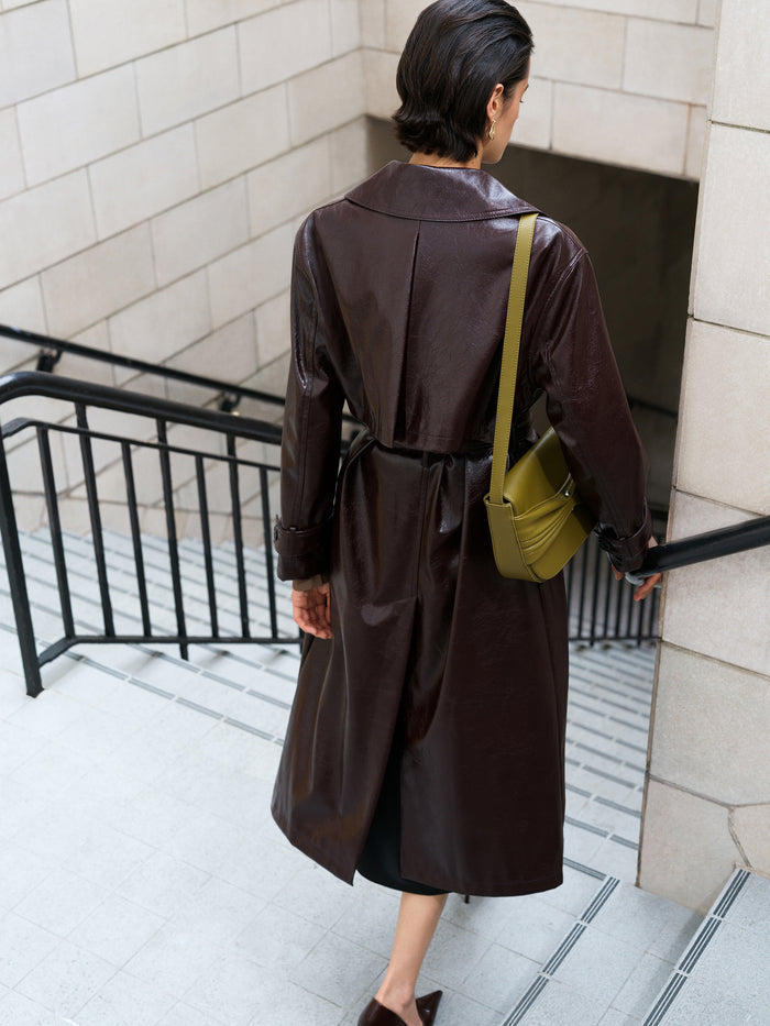 A woman wearing a burgundy belted  PU trench coat with wide lapels, side pockets and long sleeves.