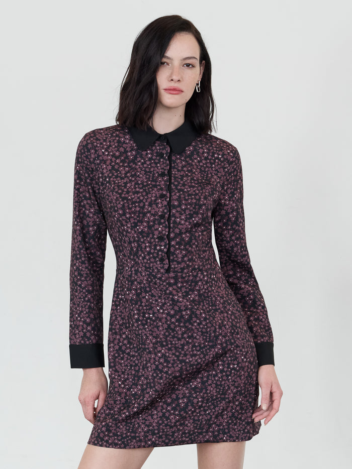 A woman wearing a black and purple floral tailored mini dress with long sleeves, rounded collar, side zip and scalloped placket.