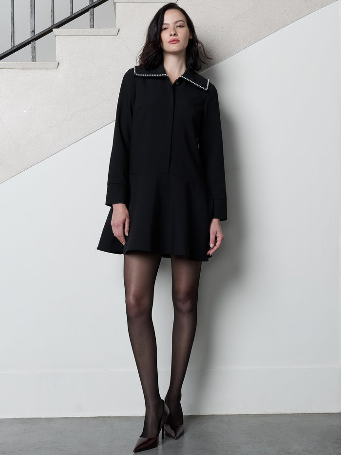 A woman wearing a black long sleeved mini dress with an oversized faux pearl collar, softly flared silhouette and a concealed front button fastening.