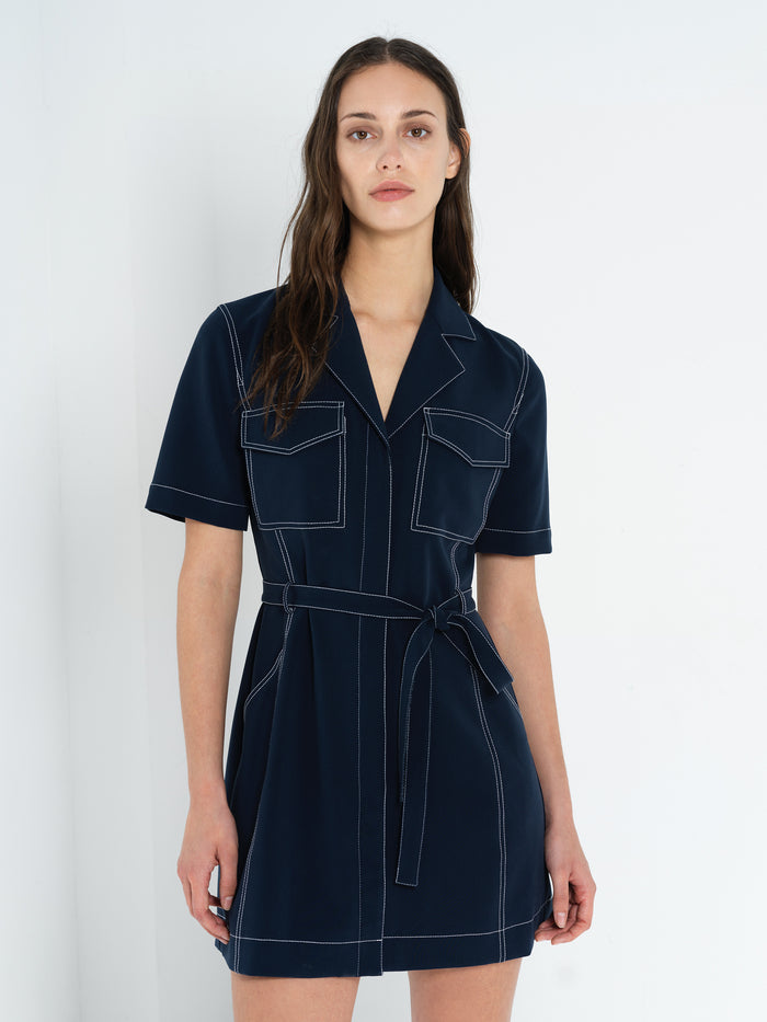 A woman wearing a belted navy blue mini dress with contrast stitching, a revere collar and button down fastening.