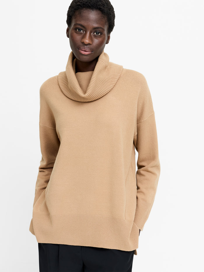 A woman wearing a beige jumper with a cowl neck, long sleeves and a relaxed fit.