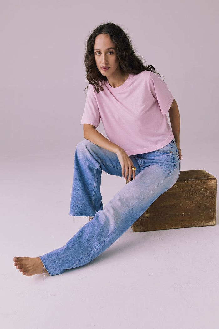 Woman sitting on an apple box wearing the Chaser Beau Nose Pink Tee styled casually with light-wash denim for a laid-back look