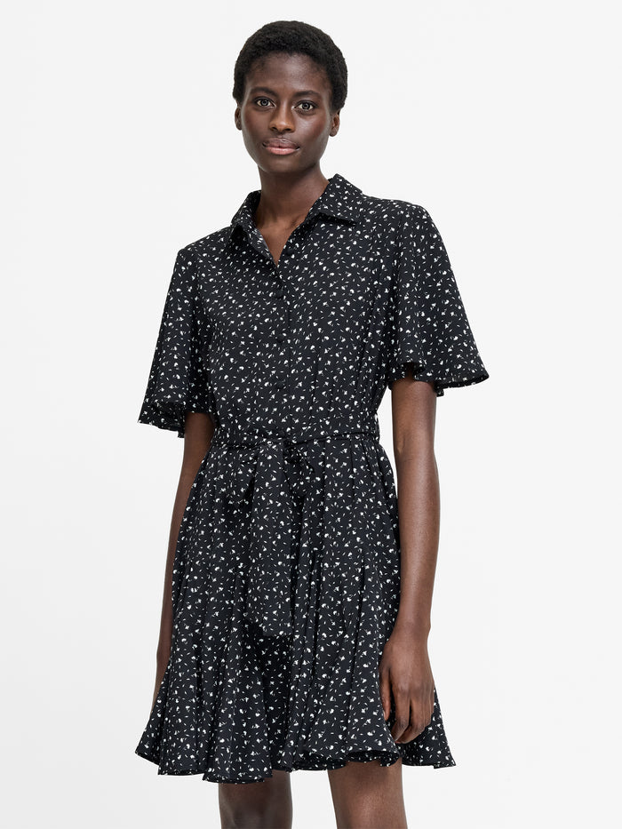 A woman wearing a black short sleeve shirt dress with a white floral pattern, button down fastening, a self tie belt and a flared hem.
