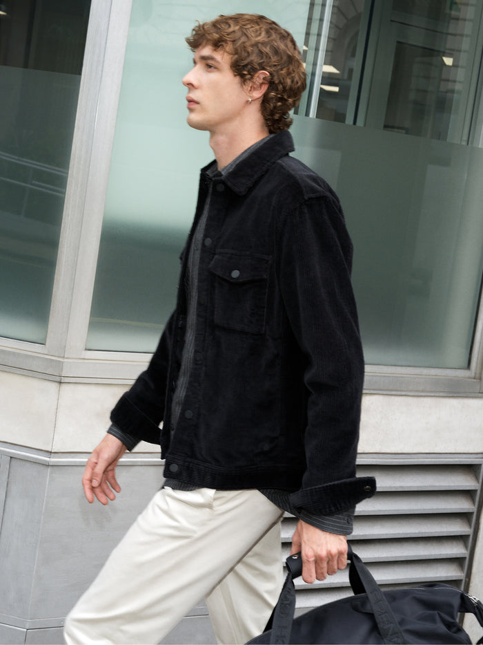 A man wearing a corduroy black Jacket with long sleeves, a point collar, 2 chested pockets, and a snap button fastening.
