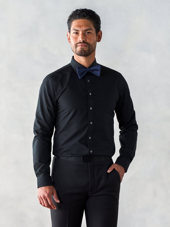 Man wearing a black shirt and blue bow tie, standing against a light background.