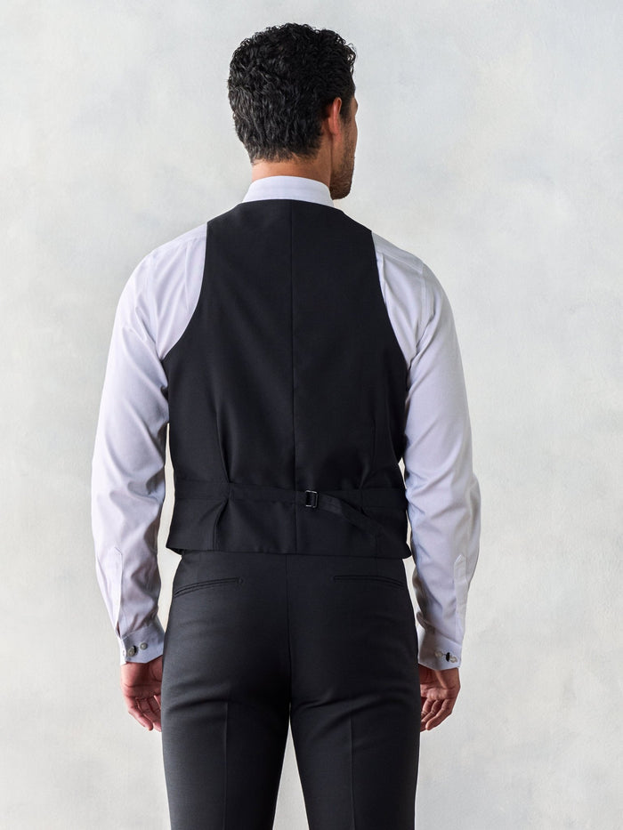 Man in a black tuxedo vest, back view, showing adjustable strap, grey backdrop.