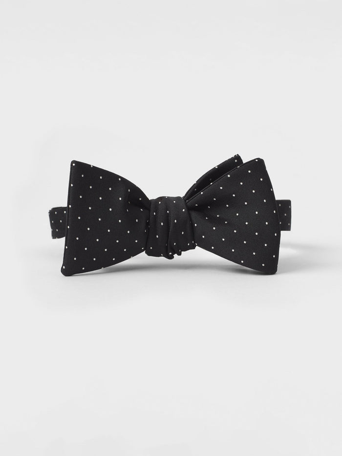 Black silk bow tie with white pindots, tied, on a grey background.