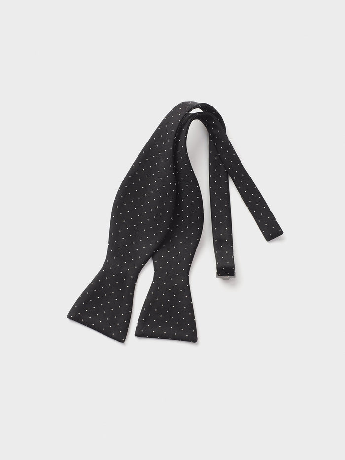 Black silk bow tie with white pindots, untied, on a light grey background.