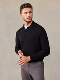 Man in a black long-sleeve polo sweater and grey trousers, beige backdrop.