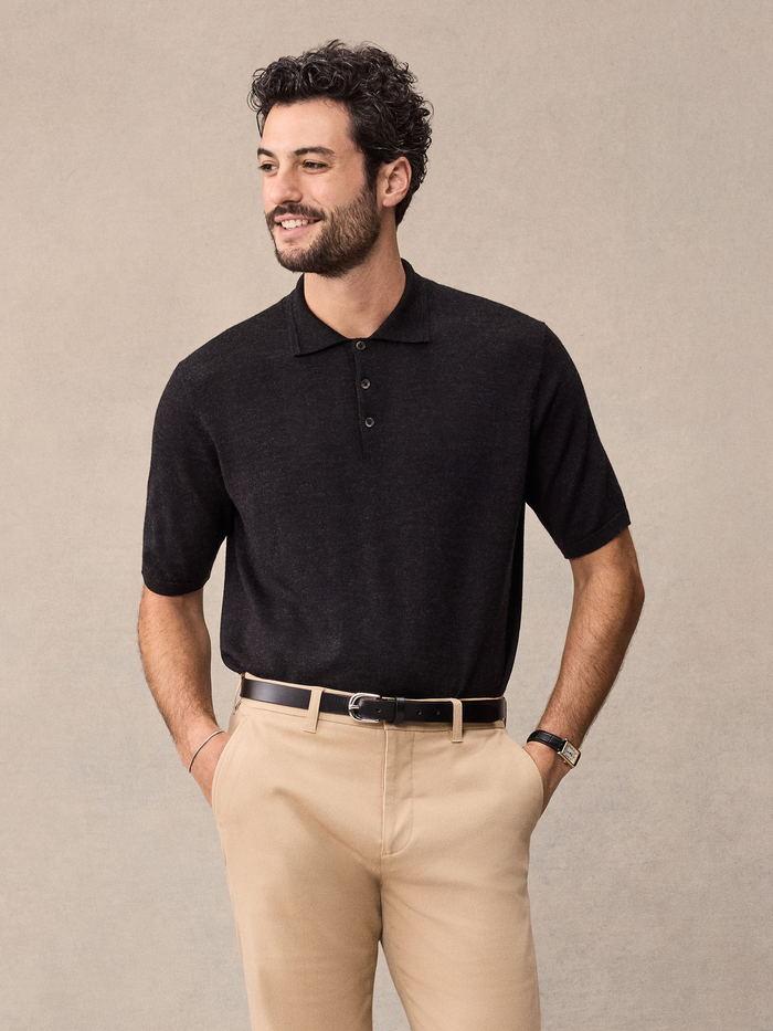 Man in a black short-sleeve sweater polo and tan trousers, beige backdrop.