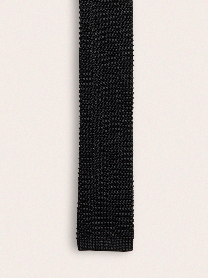 Close-up of black silk knit tie showing textured weave, cream background.