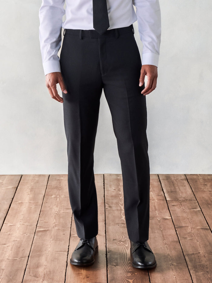 Close-up of black suit trousers worn with white shirt and black tie, wood floor.