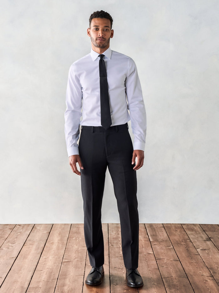 Man in a white dress shirt, black tie, and black suit trousers, wood floor backdrop.