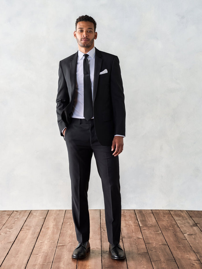 Man in a black suit with tie and pocket square, full length, wood floor backdrop.
