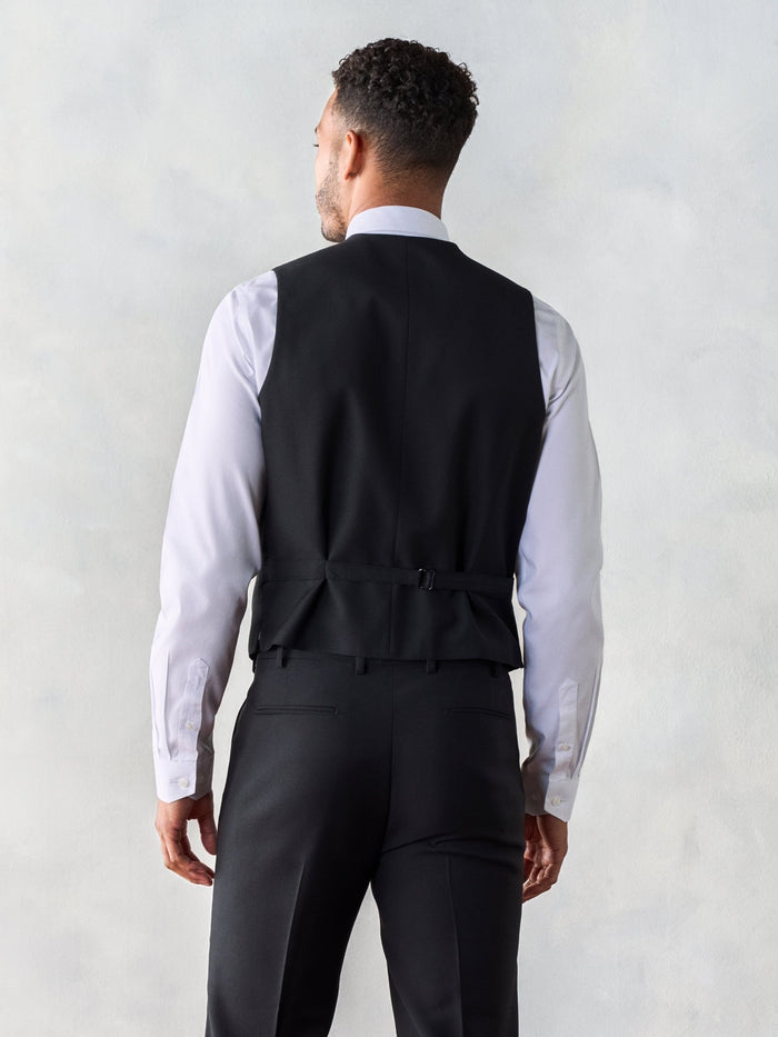 Man in a black suit vest, back view, showing adjustable strap, grey backdrop.
