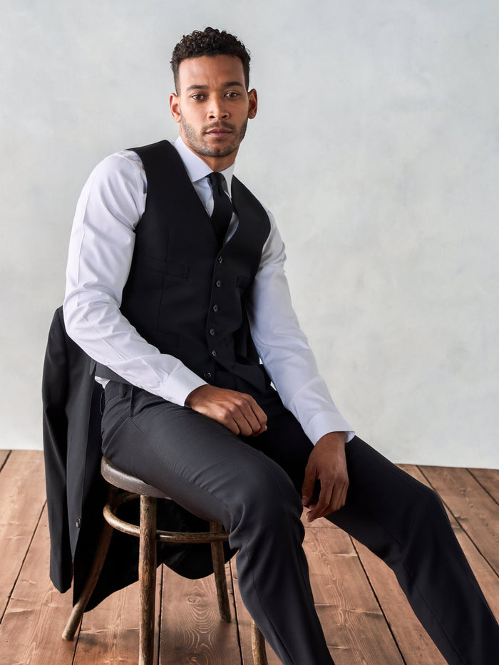 Man in a black suit vest seated, jacket draped over a stool, wood floor backdrop.
