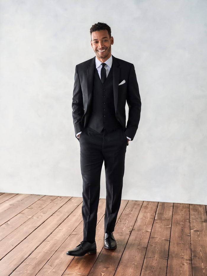 Man in a black suit vest, full-length front view, smiling, wood floor backdrop.