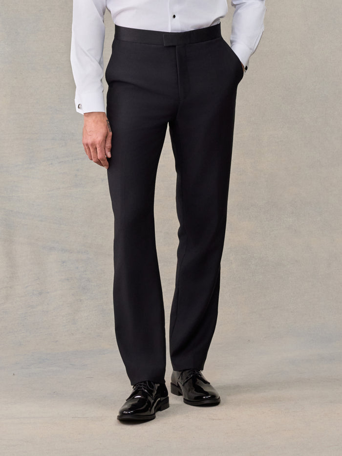 Close-up of black slim tuxedo trousers worn with white shirt, beige backdrop.