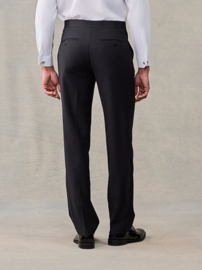 Close-up back view of black slim tuxedo trousers, white shirt, beige backdrop.