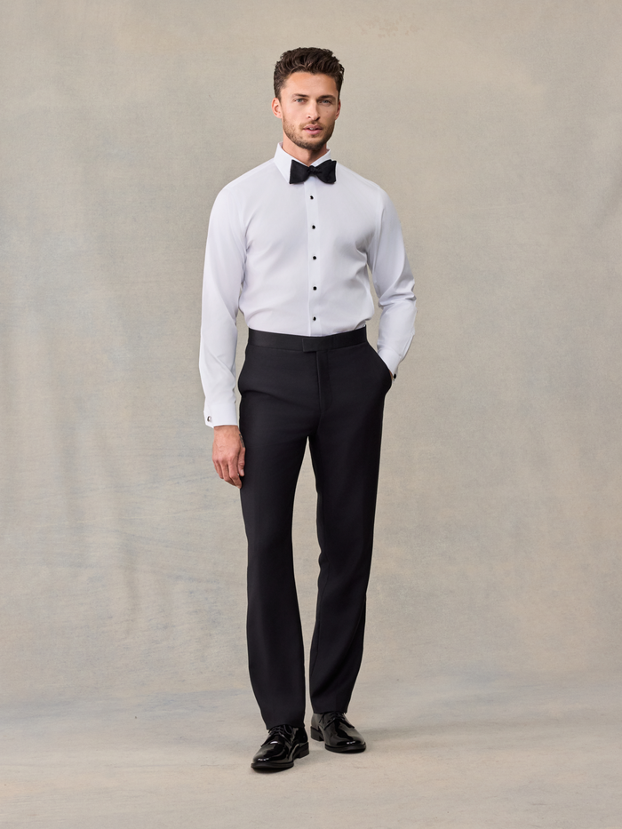 Man in white tuxedo shirt and black trousers with black bow tie, front view, on beige backdrop