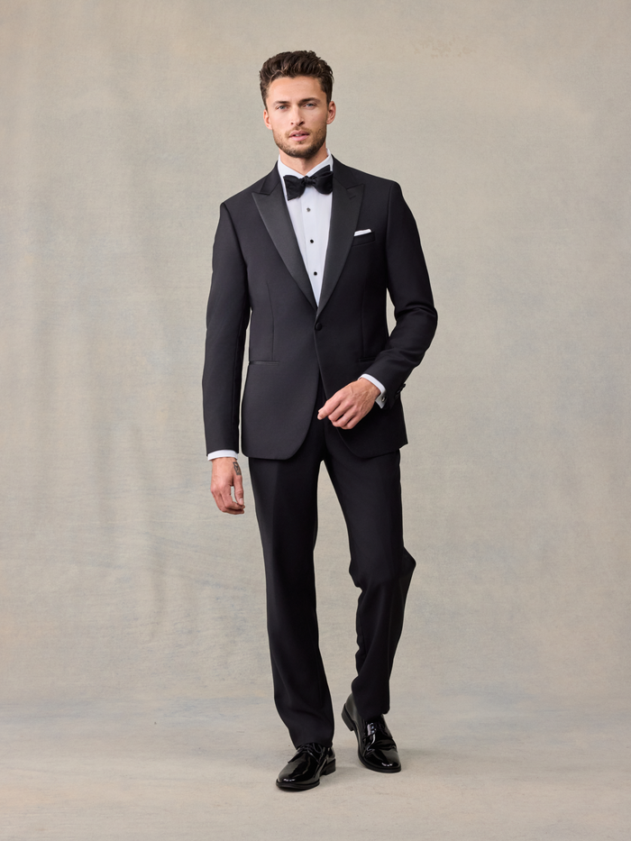 Man in black peak lapel tuxedo with bow tie and white pocket square, walking, on beige backdrop