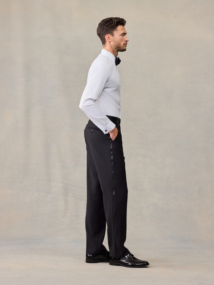 Man in white tuxedo shirt and black trousers with black bow tie, side profile, on beige backdrop