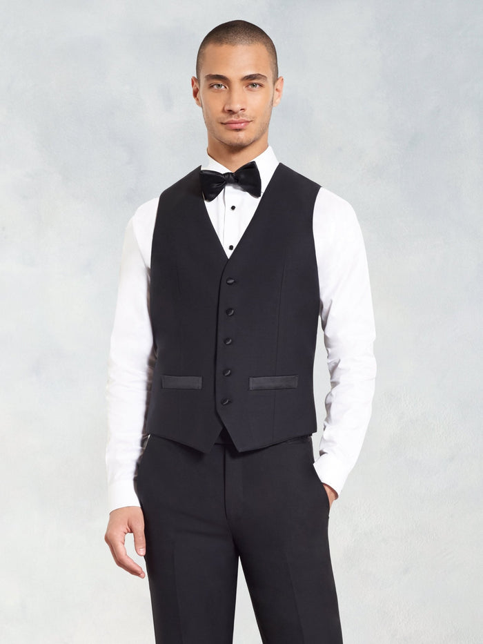 Man in a black tuxedo vest with black bow tie and white shirt, grey backdrop.