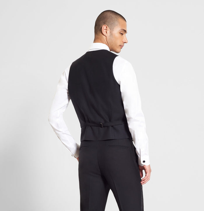 Man in a black tuxedo vest, back view, showing adjustable strap, grey backdrop.