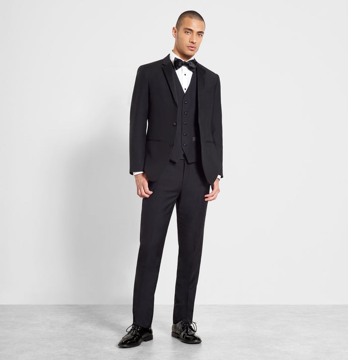 Man in a black tuxedo vest and jacket with black bow tie, full length, grey backdrop.