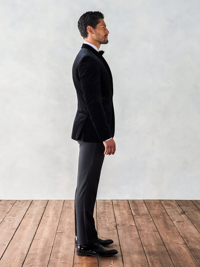 Man in a black velvet tuxedo jacket, side profile, full length, wood floor backdrop.