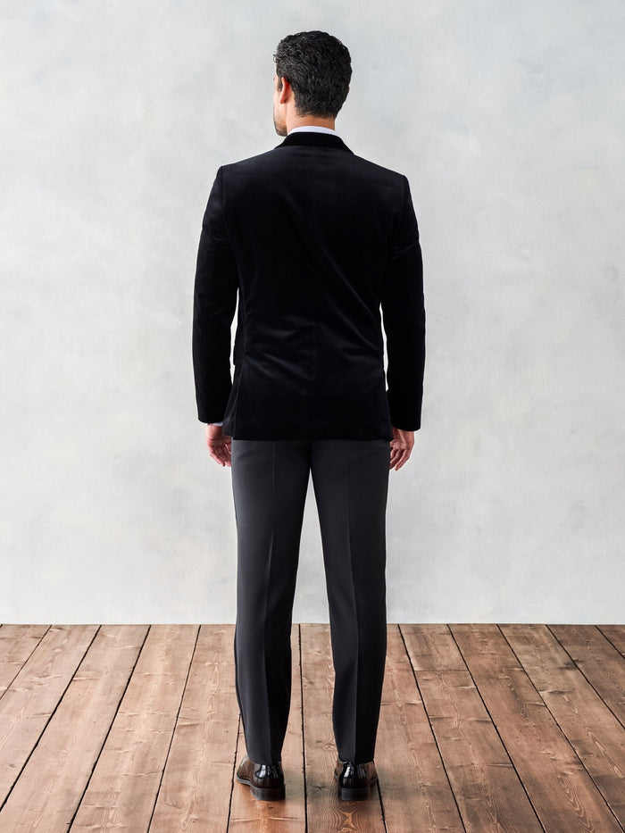 Man in a black velvet tuxedo jacket, back view, full length, wood floor backdrop.