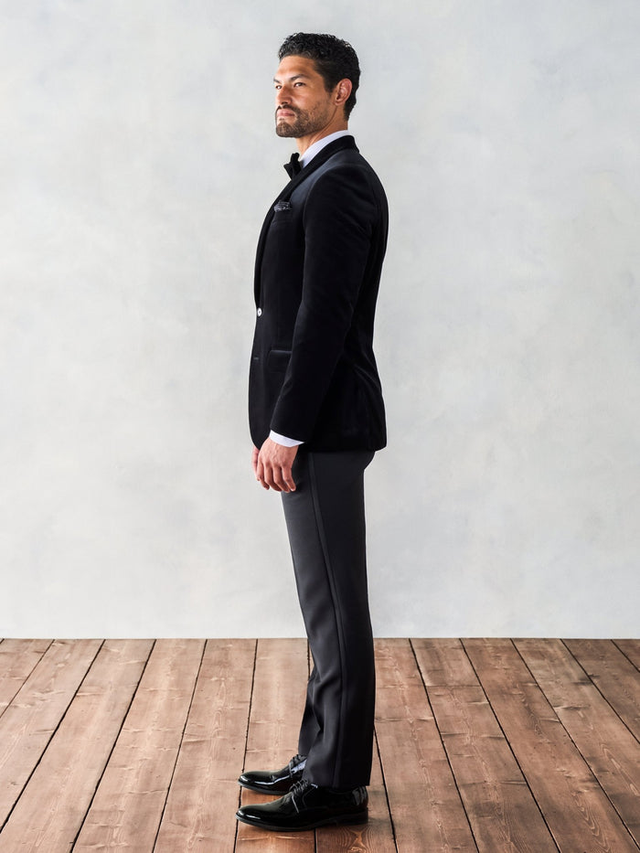 Man in a black velvet tuxedo jacket, side profile, full length, wood floor backdrop.