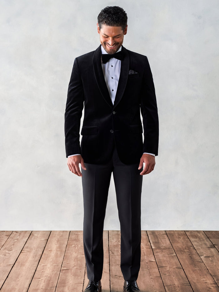 Man in a black velvet tuxedo jacket, smiling, full length, wood floor backdrop.