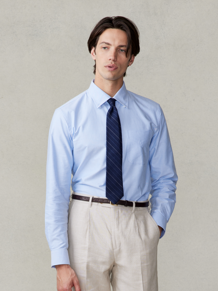 Cropped front view of a man in a light blue oxford shirt, navy striped tie, and tan trousers.