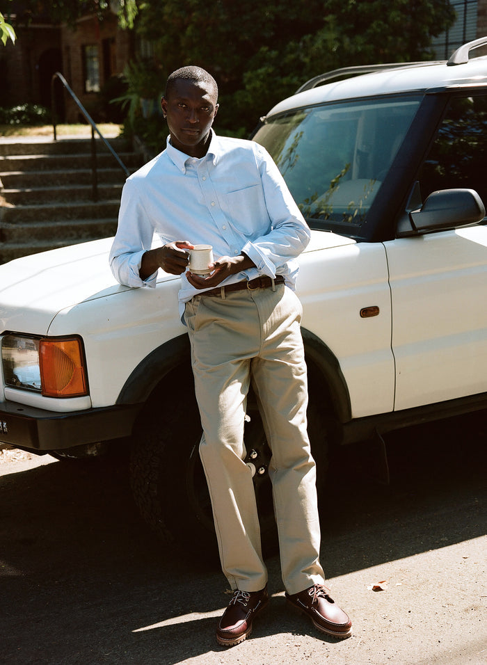 Man in a light blue oxford shirt and tan chinos leaning against a white SUV outdoors.