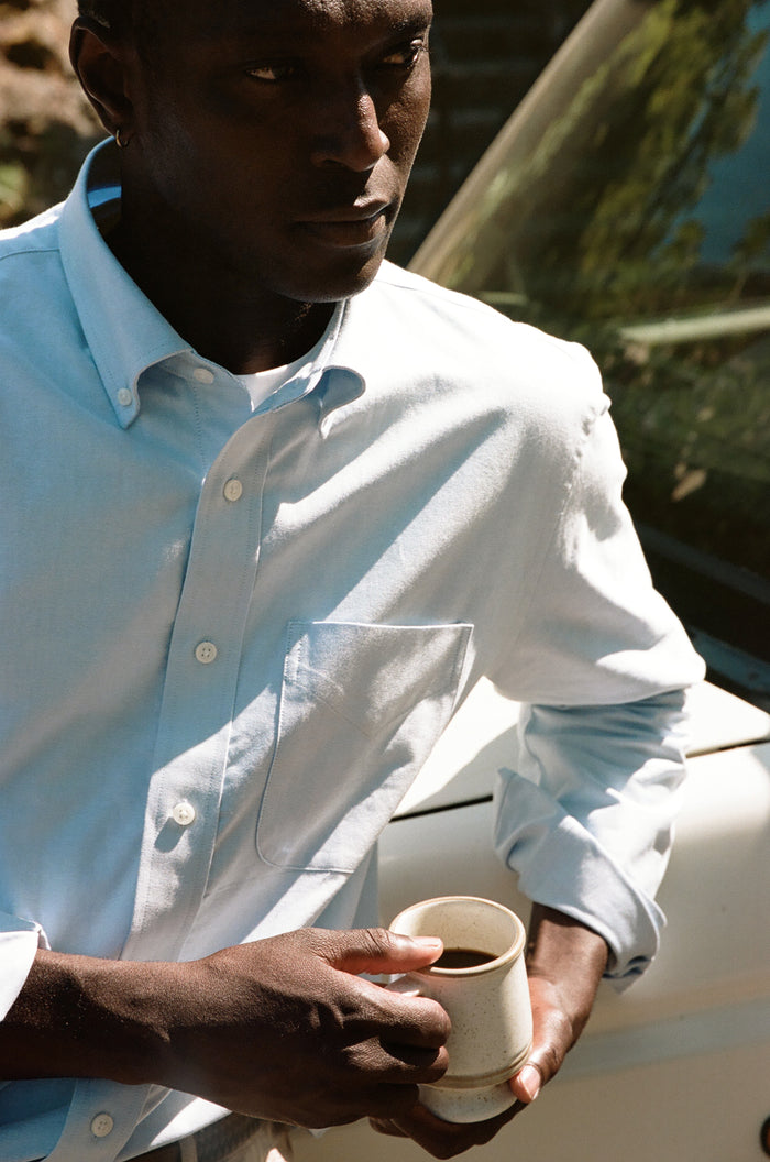 Close-up of a man in a light blue oxford shirt holding a coffee mug outdoors.