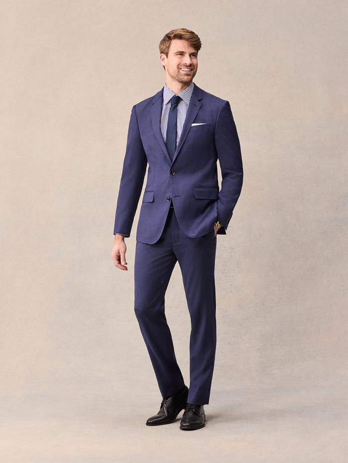 Man in a blue suit with striped shirt and navy tie, full length on a beige backdrop